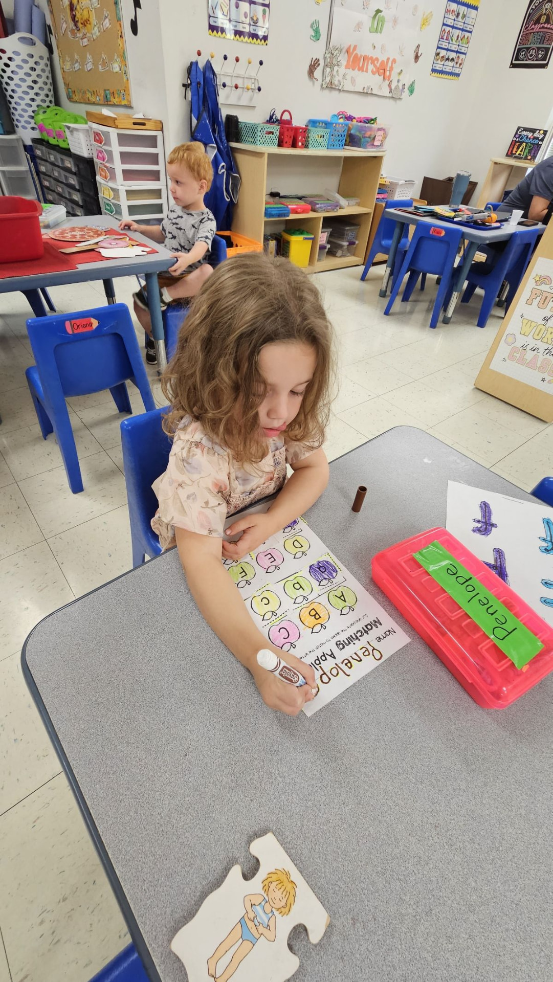 A preschool girl matching letters on a worksheet at a classroom table.
