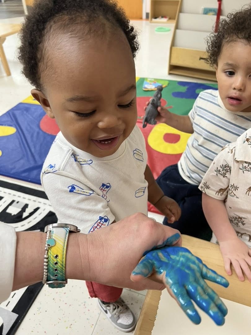 A grinning toddler holding out a hand a teacher has just painted blue, while a friend watches.