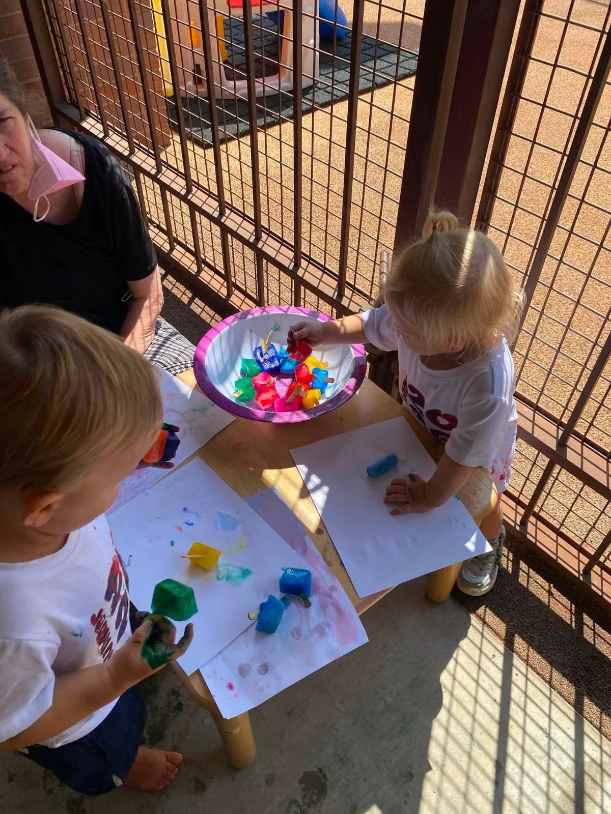 Toddlers painting with colored ice cubes on a shaded porch.