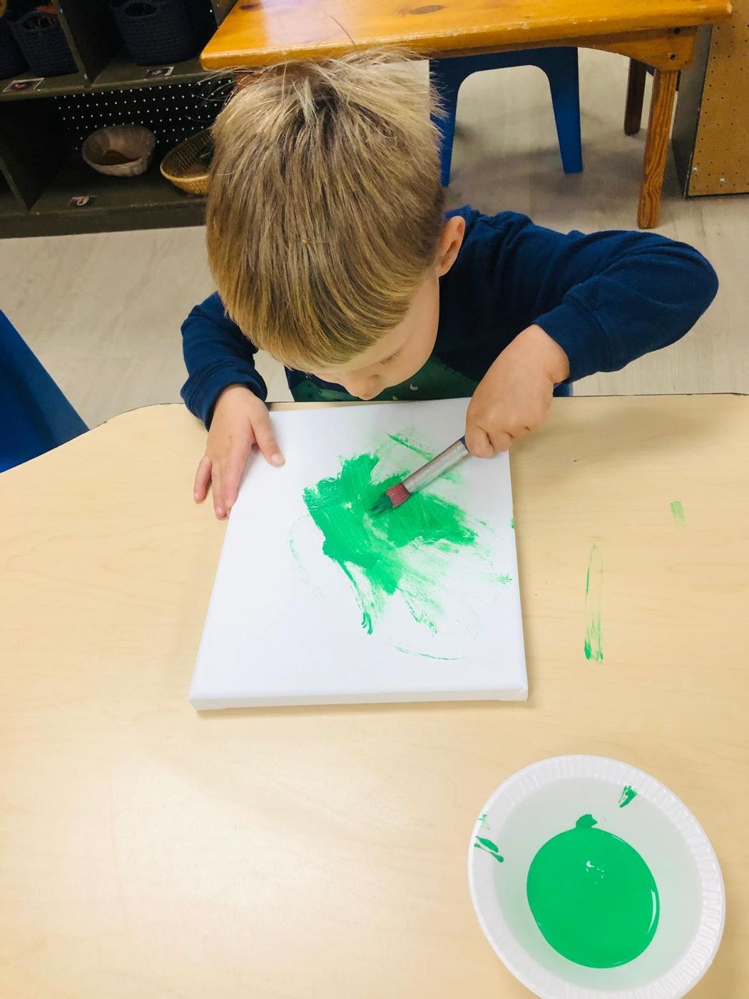 A young boy paints a small canvas bright green with a brush at his table.