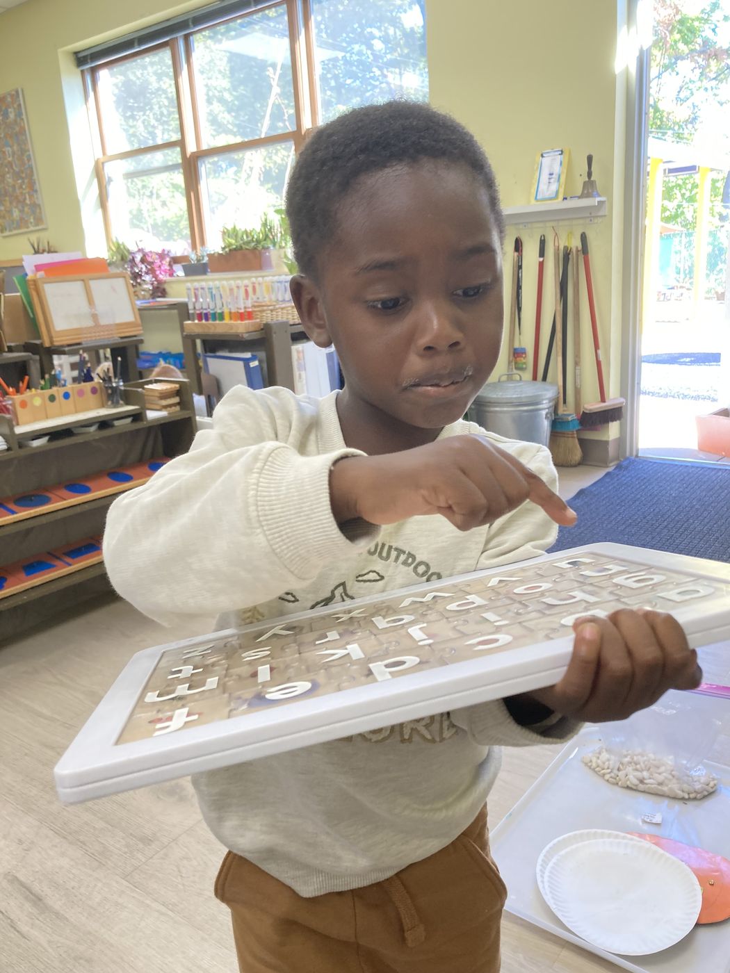 A boy holds up a wooden tray of lowercase letters, pointing to one with his finger.