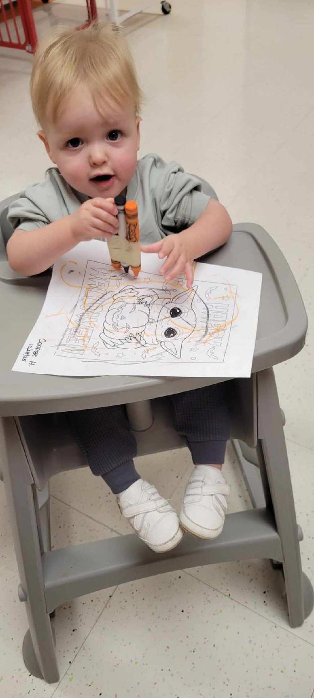 A baby in a high chair holds a crayon over a coloring page and looks up.