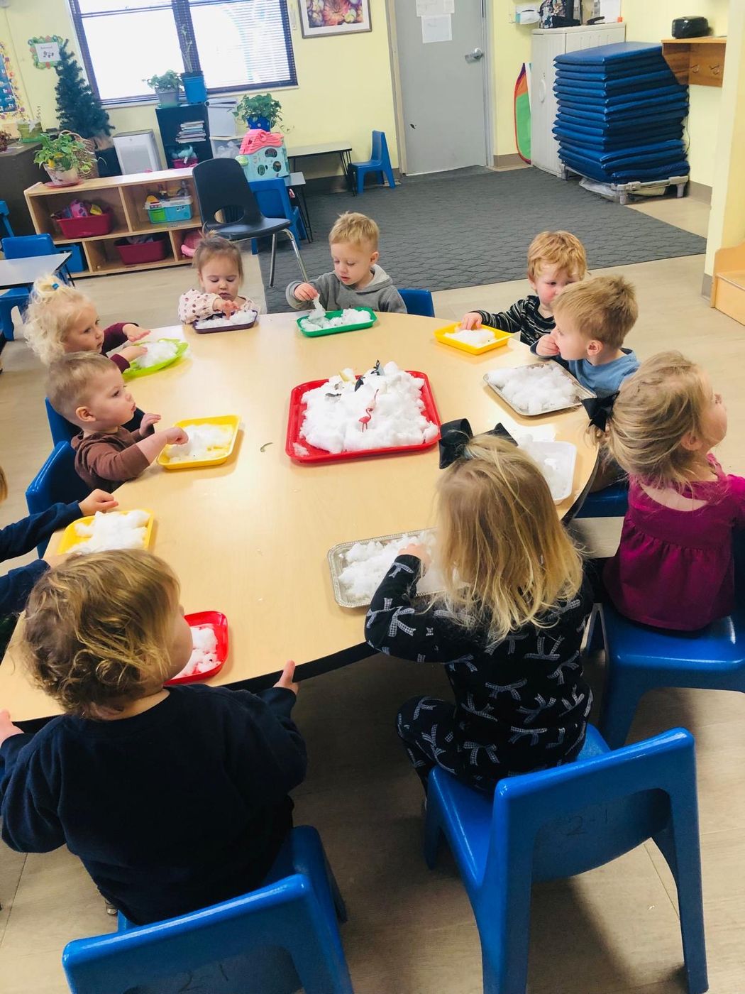Toddlers sit around a round table exploring trays of fluffy white snow-like material.