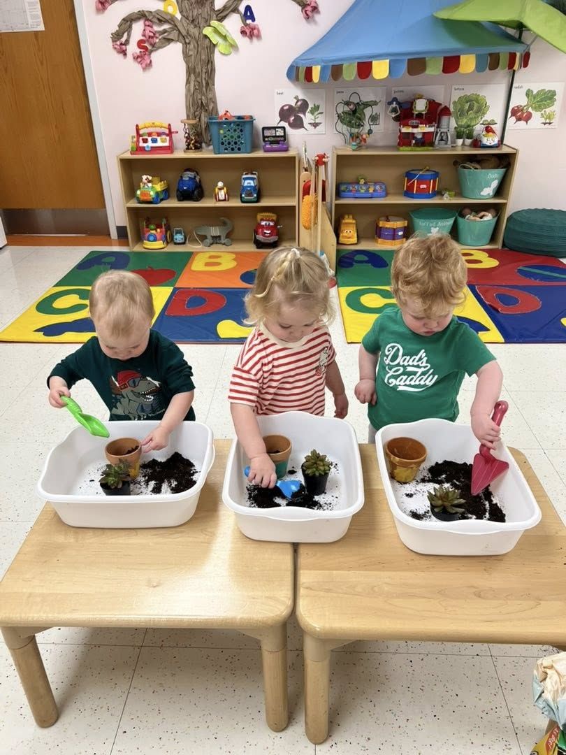Three toddlers scoop soil into small pots in bins at a low table.