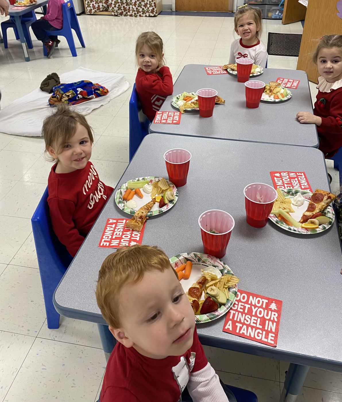 Children in holiday sweaters sit at classroom tables with plates of party food and red cups.