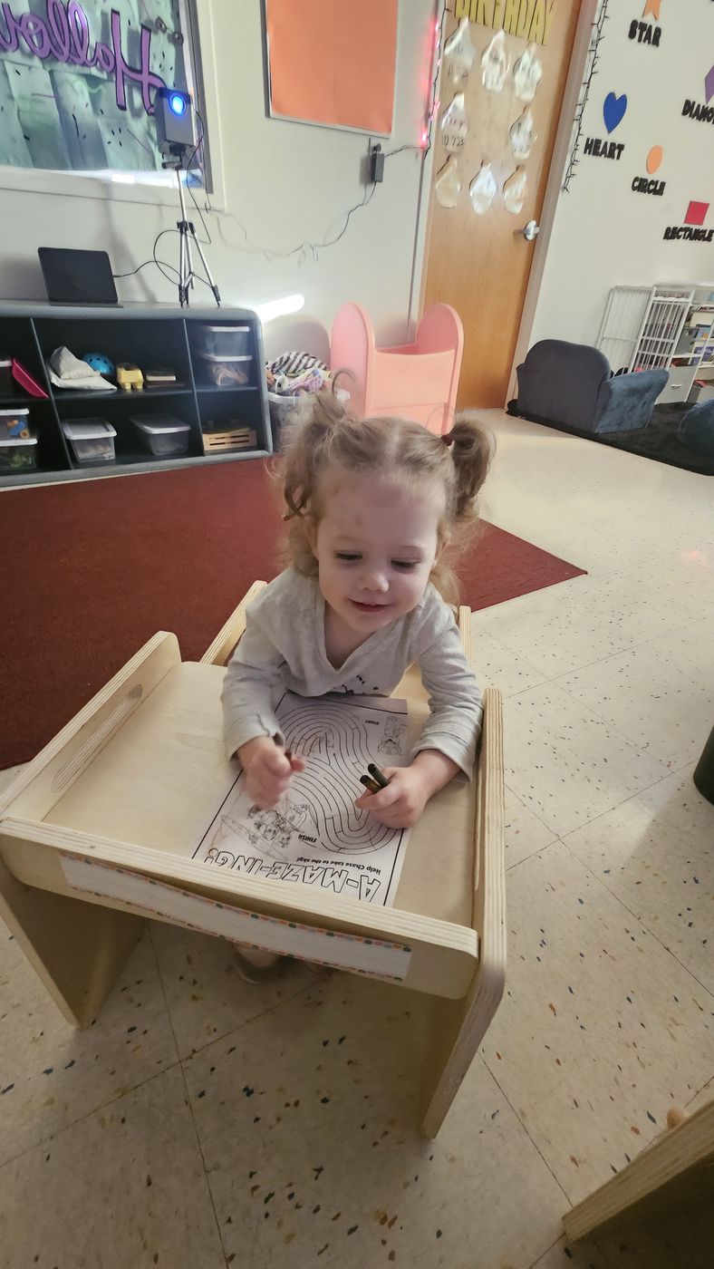 A toddler with pigtails colors a worksheet at a small wooden desk.