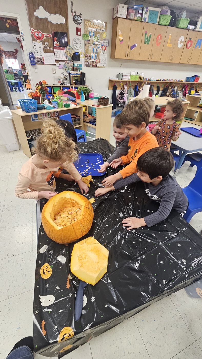 Children gather around a table covered in black plastic to scoop out a carved pumpkin.