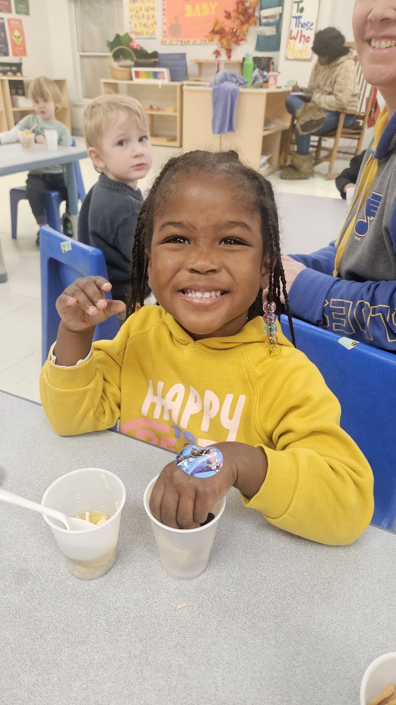 A smiling girl in a yellow sweatshirt holds a snack cup at her classroom table.