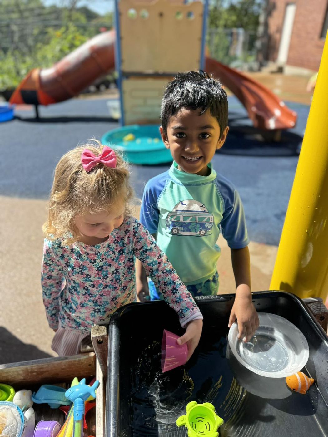 Two children play with cups and scoops at an outdoor water table on a sunny day.