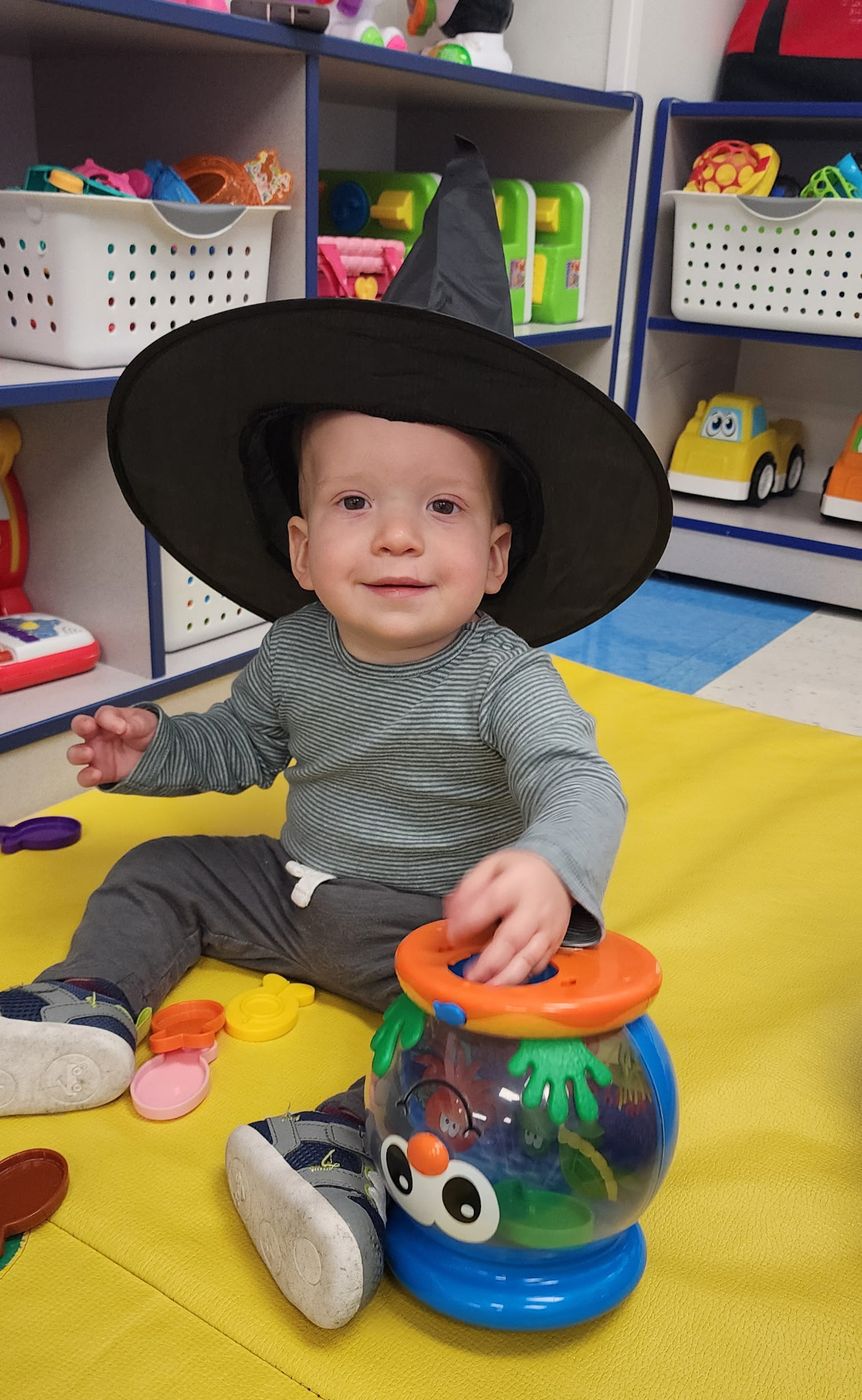 A smiling baby in a black witch hat sits on a yellow mat playing with a toy fishbowl.
