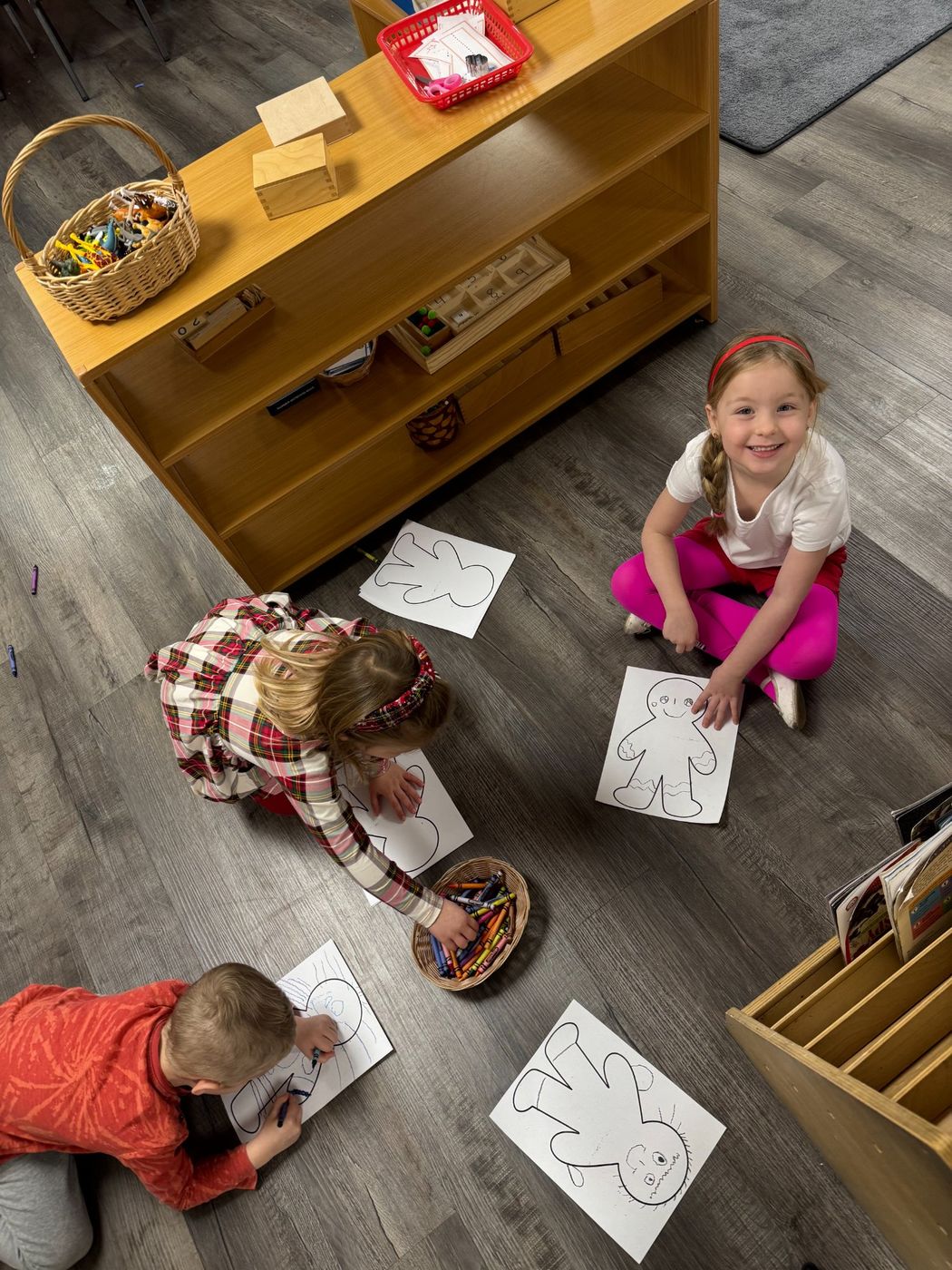 Three children lie on the classroom floor coloring gingerbread outlines on paper.