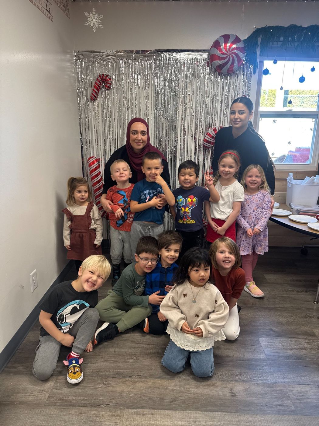 A group of children and two teachers pose together in front of a sparkly holiday backdrop.