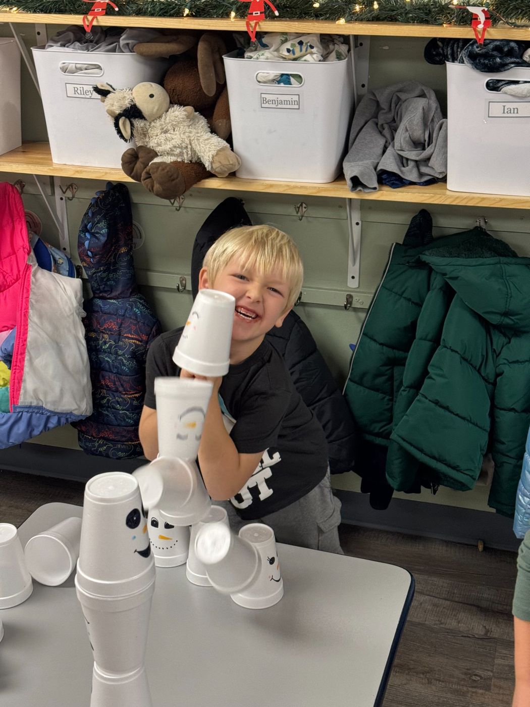 A laughing boy holds a stack of paper cups up to his face beside a cubby wall.