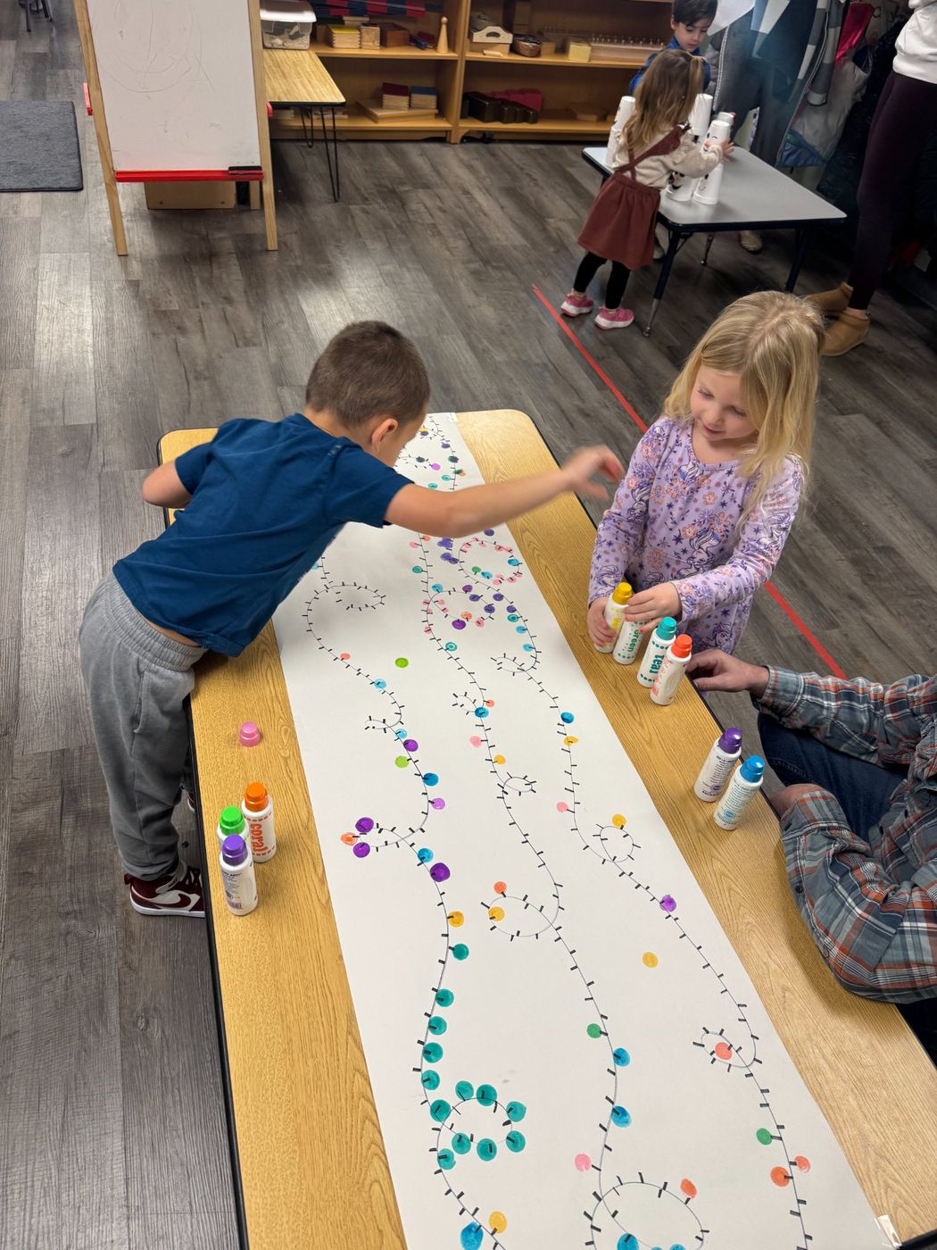 Children dab colorful dot markers along a long paper banner on a table.