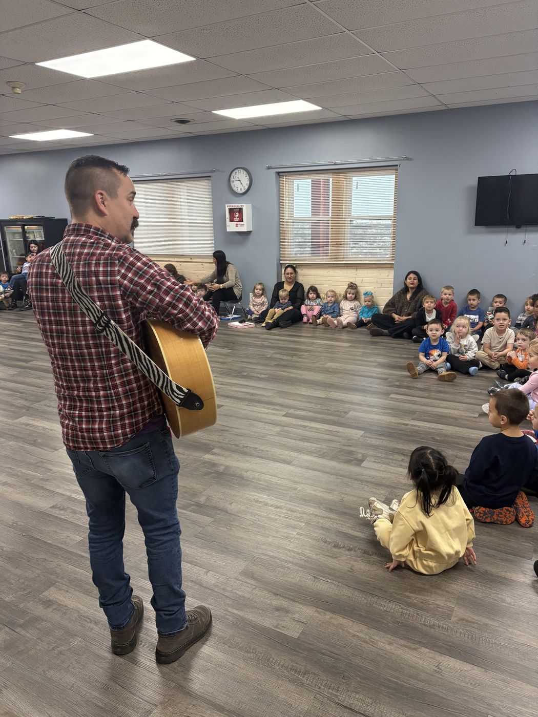 A man plays guitar for a large group of children seated on the floor of a big room.