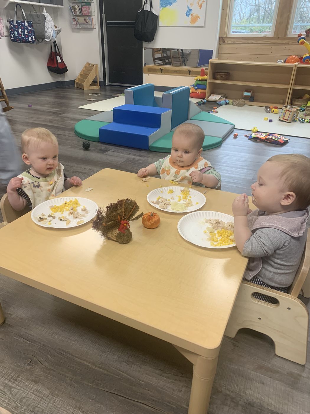 Infants sit in low wooden chairs around a small table eating a festive lunch.