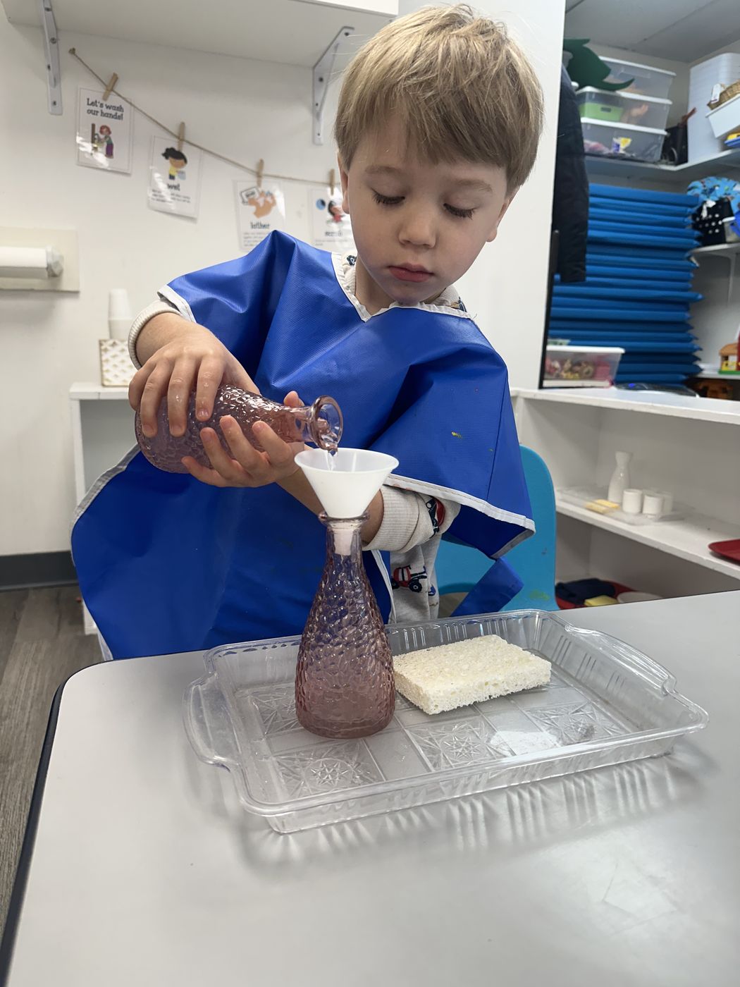 A boy in a blue smock pours liquid through a funnel into a glass bottle on a tray.