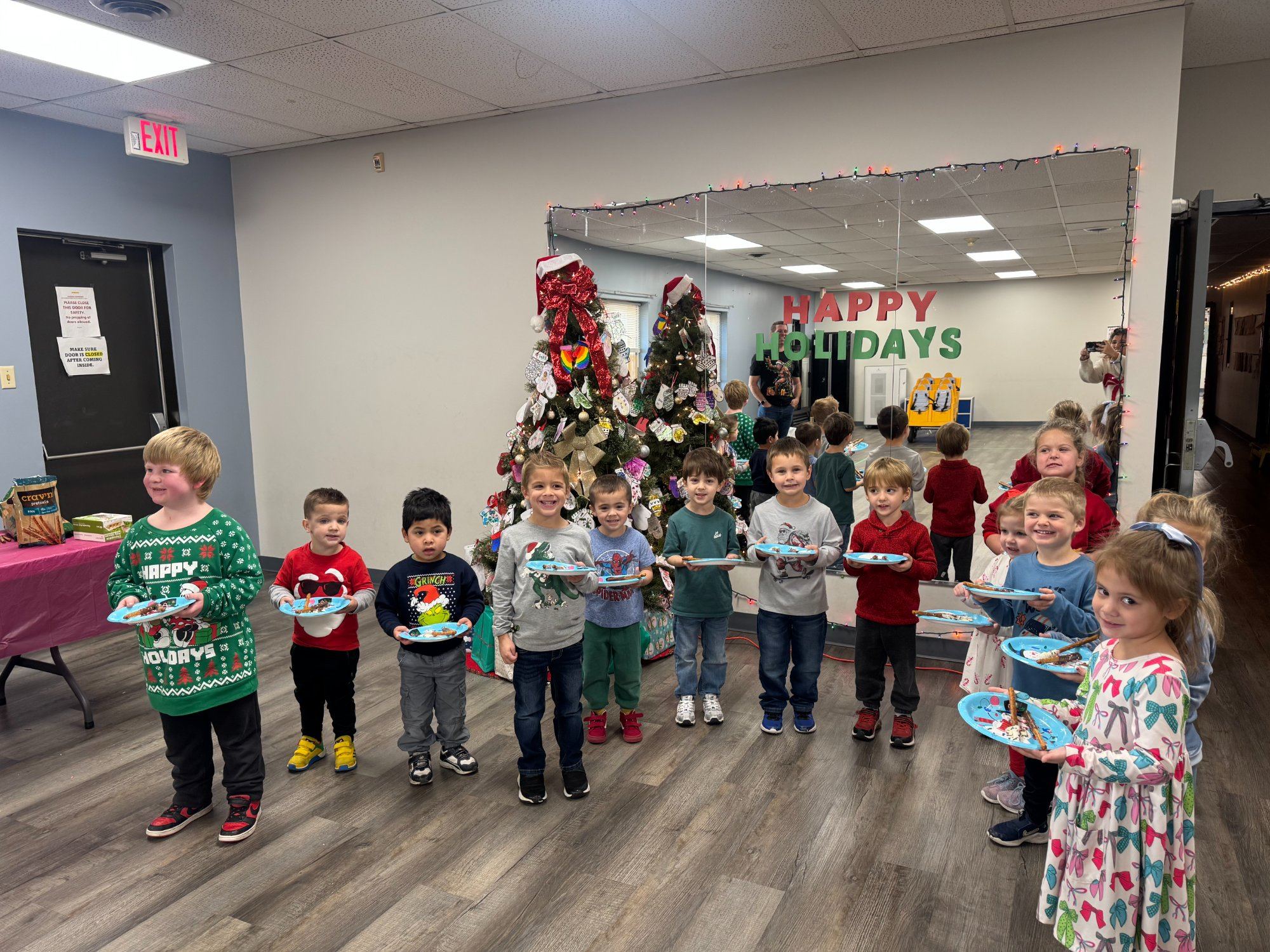A line of children holding decorated plates stands in front of holiday trees in a big room.