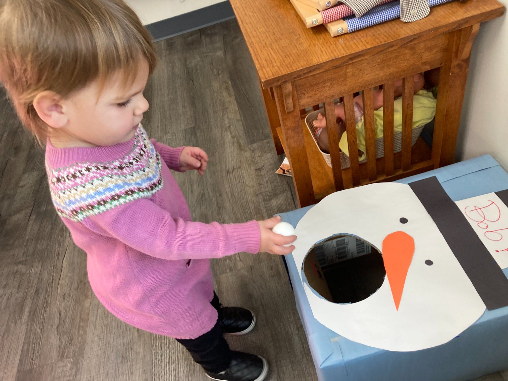A toddler girl in a pink sweater drops a ball into a snowman box during a holiday game.