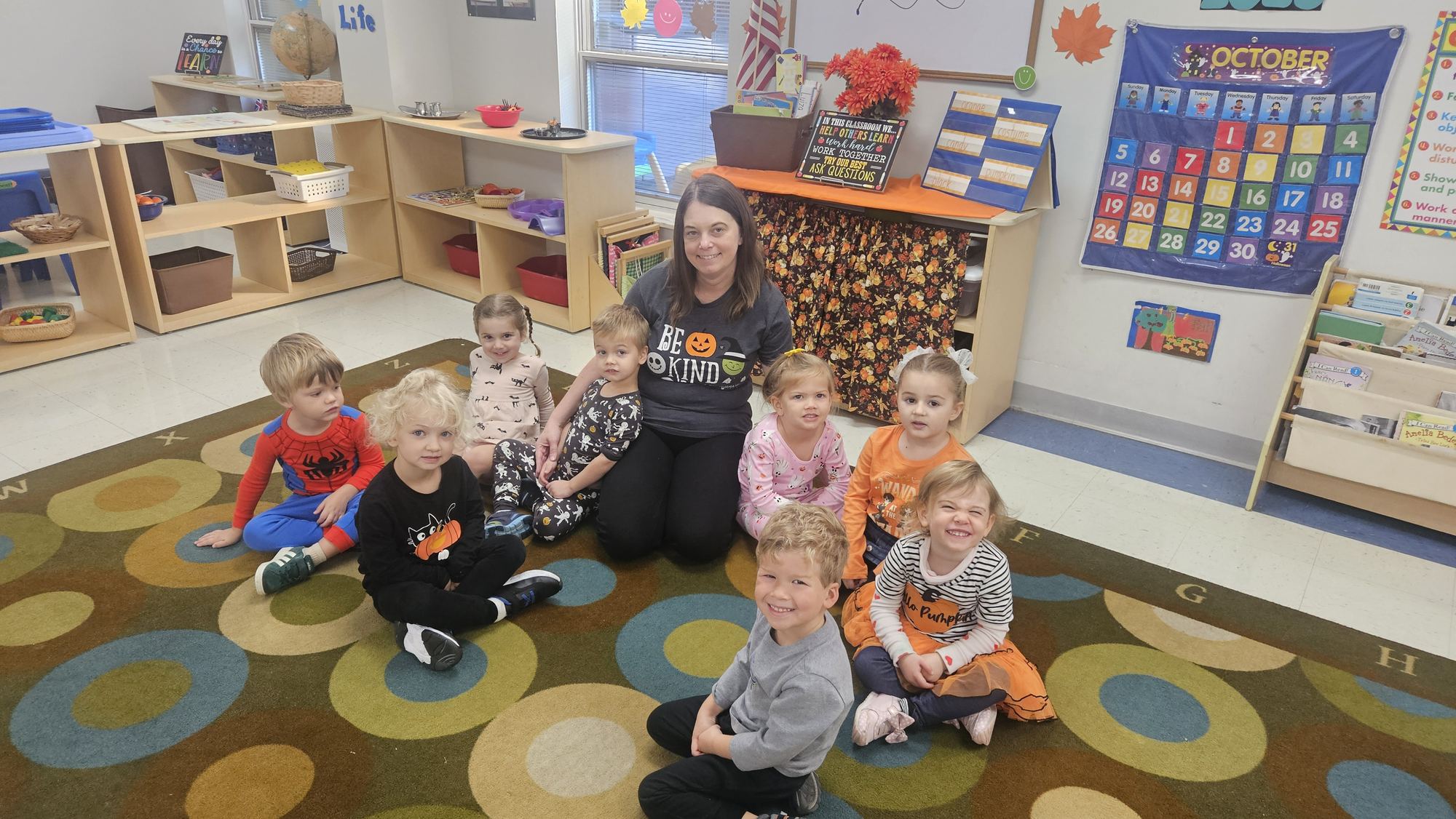 A teacher sits on a patterned rug with a group of toddlers in Halloween shirts.