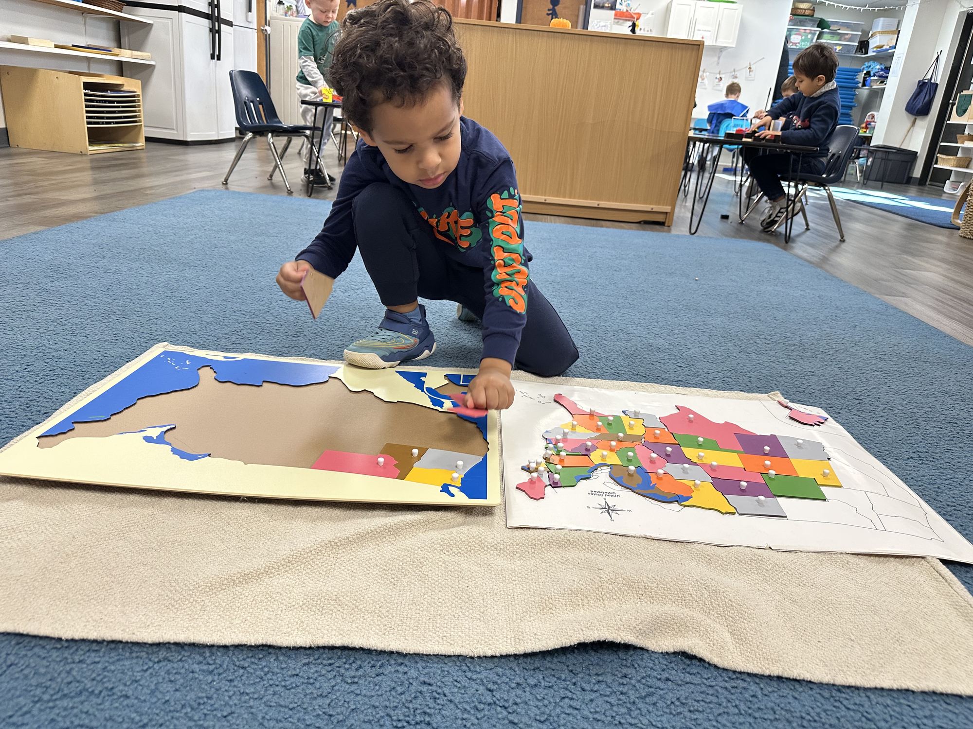 A boy kneels on a rug fitting pieces into a wooden puzzle map of the United States.