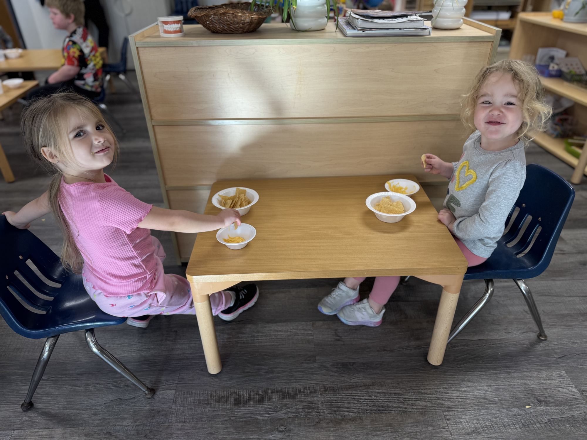 Two smiling girls eat a snack together at a small wooden table in a Montessori classroom.