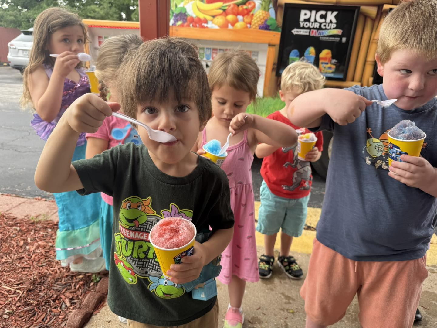Children eat snow cones with spoons outdoors on a summer day.