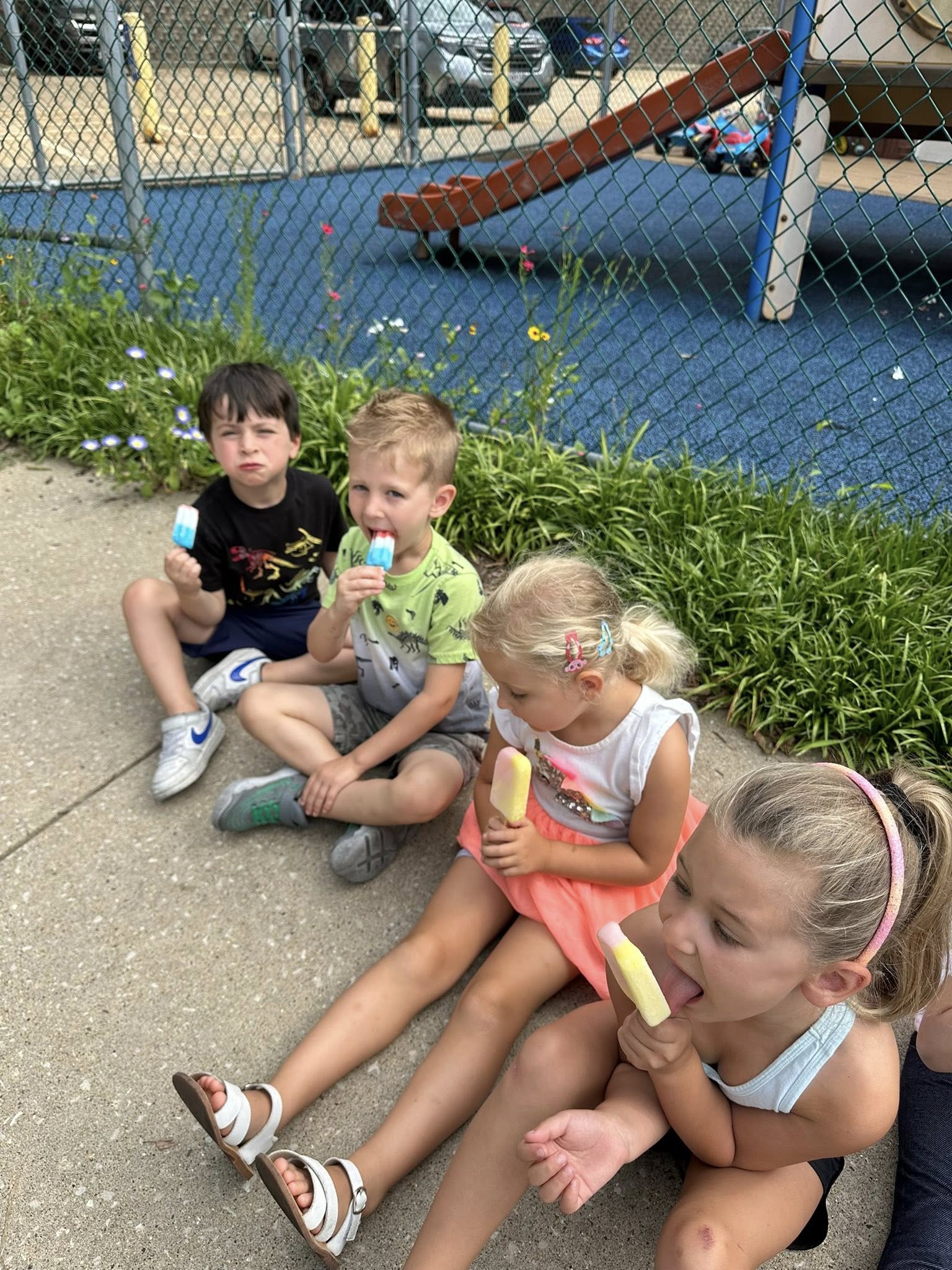 Four preschoolers eating ice lollies together beside the playground.