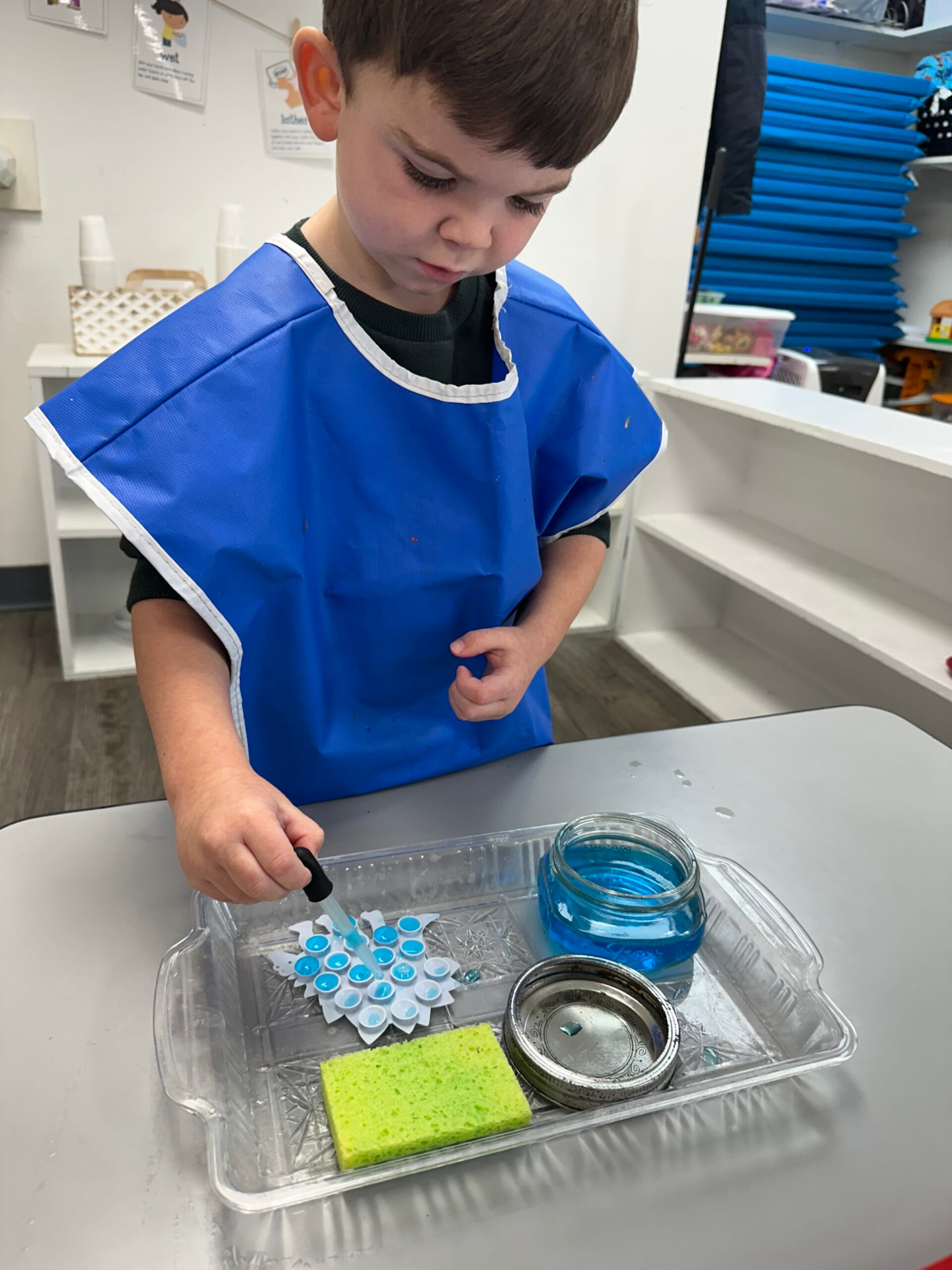 A boy in a blue smock using a dropper to fill a snowflake tray with blue water.