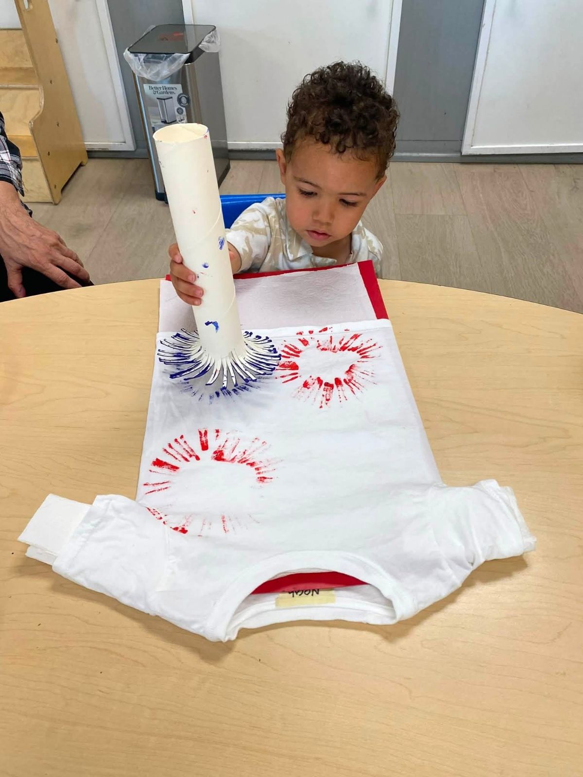 A toddler printing red and blue starbursts onto a t-shirt with a paper tube.