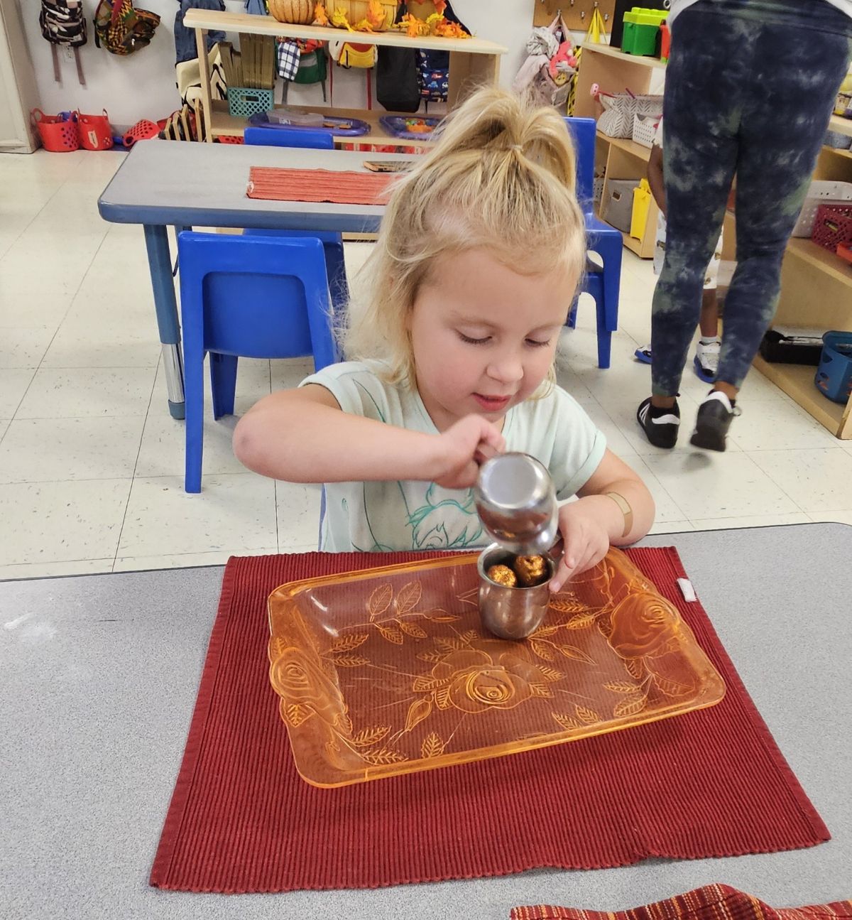 A preschool girl carefully pouring from a small metal pitcher onto a work mat.